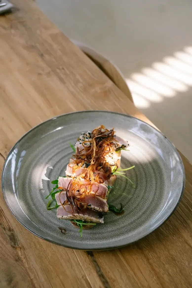 Somewhere Restaurant & Bar, Asia — Seared duck breast with crispy skin, microgreens, and caramelized garnish on slate-grey ceramic plate, natural daylight on wooden table.