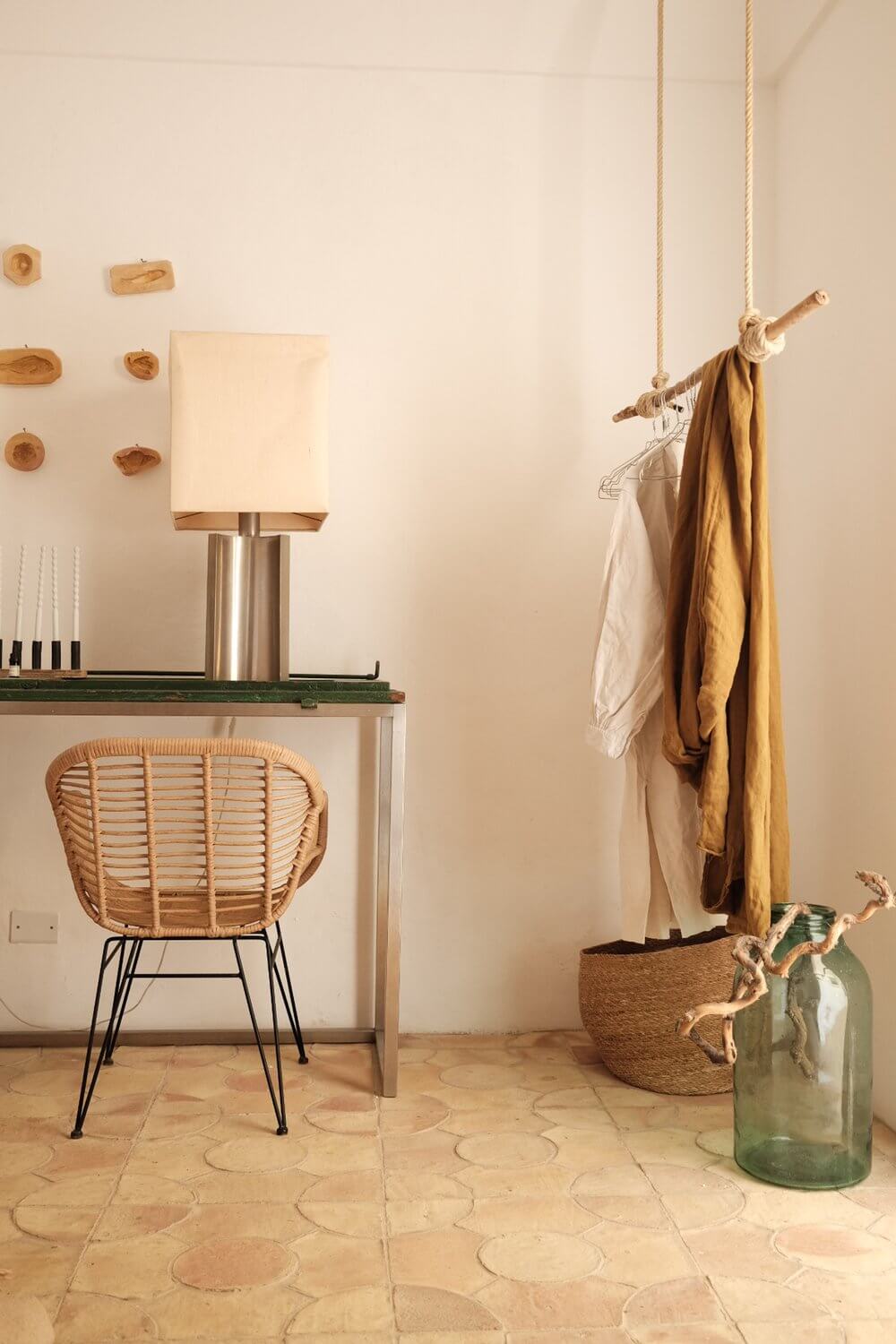 Parco Dei Sesi, Europe — Minimalist bedroom with woven rattan chair, wooden wall art, rope-hung clothing rail, terracotta tile floor, and natural linen textiles.