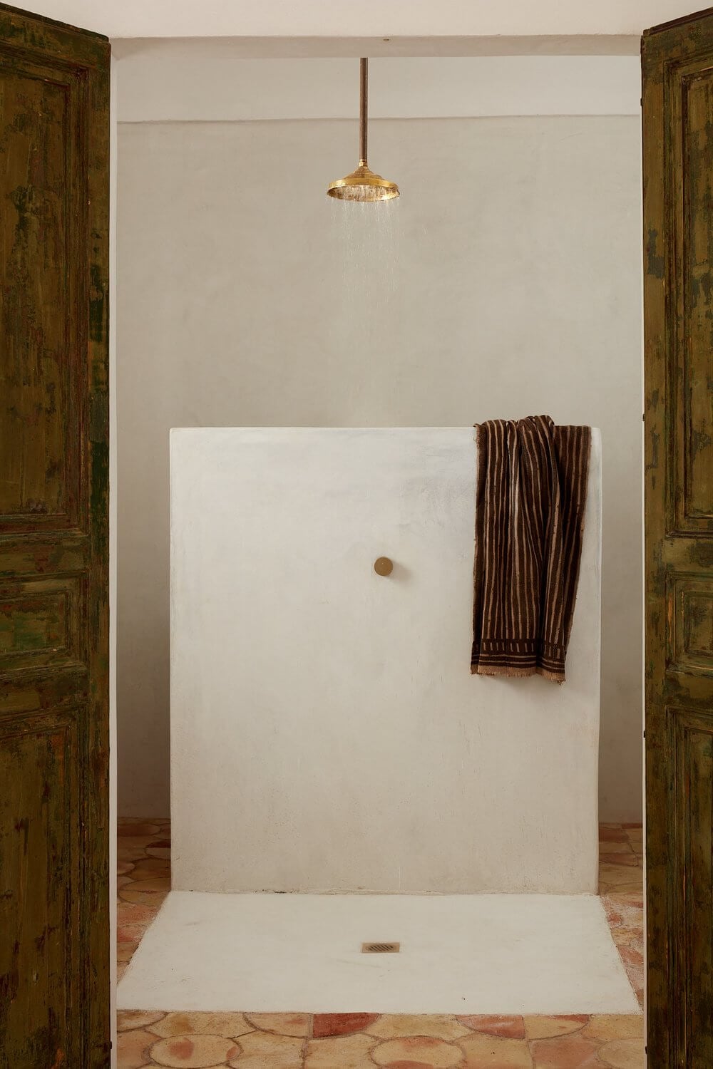 Parco Dei Sesi, Europe — Minimalist shower alcove with white plaster walls, brass rainfall showerhead, wooden doors, and striped brown towel.