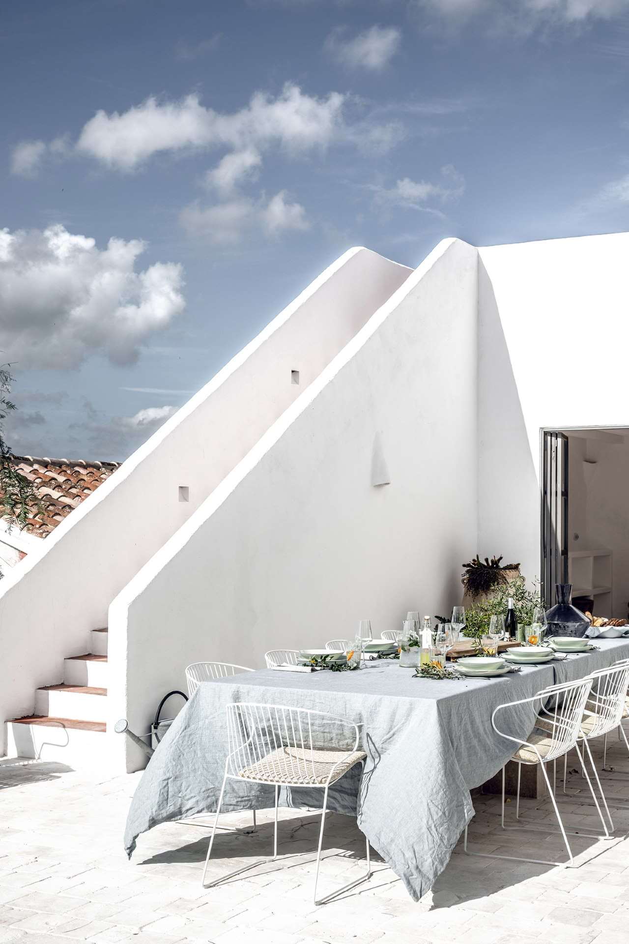 Es Bec d’Aguila — Es Bec d'Aguila, Balearic Islands, Spain — Whitewashed modernist villa terrace with dining table, wire chairs, and geometric roofline against blue sky.