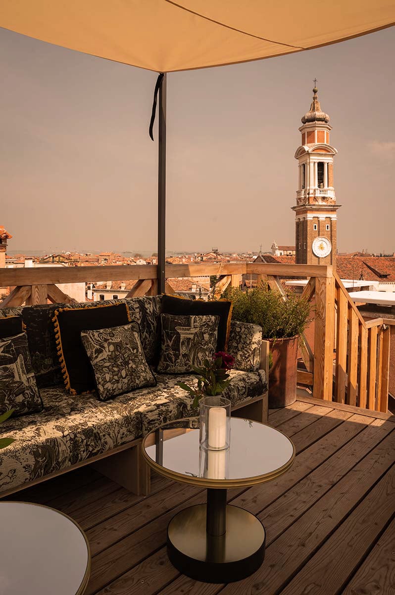 The Venice Venice Hotel, Venice, Italy — Ochre canvas canopy shading a wooden deck with patterned cushioned seating, clock tower and terracotta rooftops beyond.