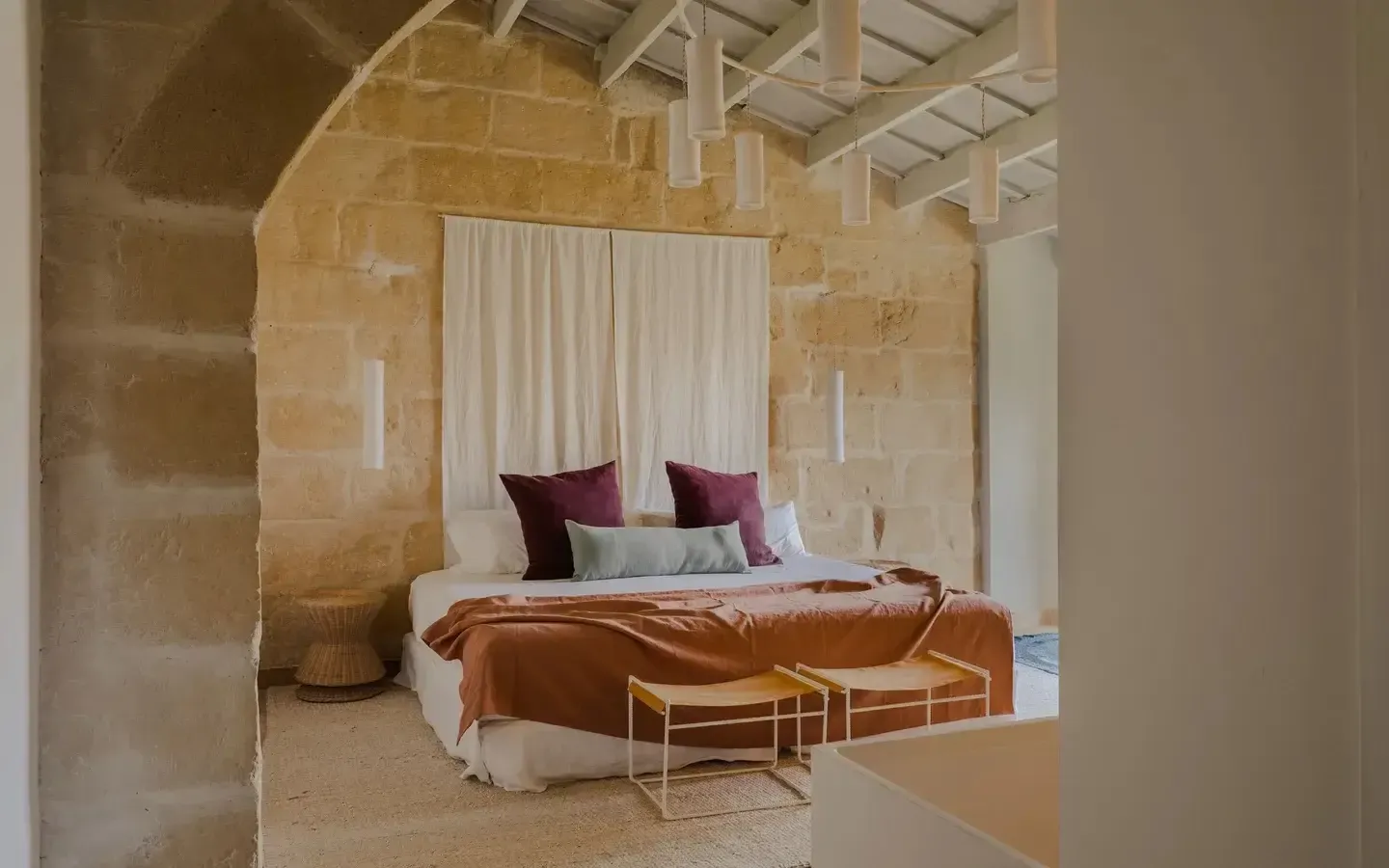 Amagatay, Balearic Islands — Canopied bed with linen curtains in stone-walled bedroom with terracotta throw and brass side tables, minimalist luxury design.