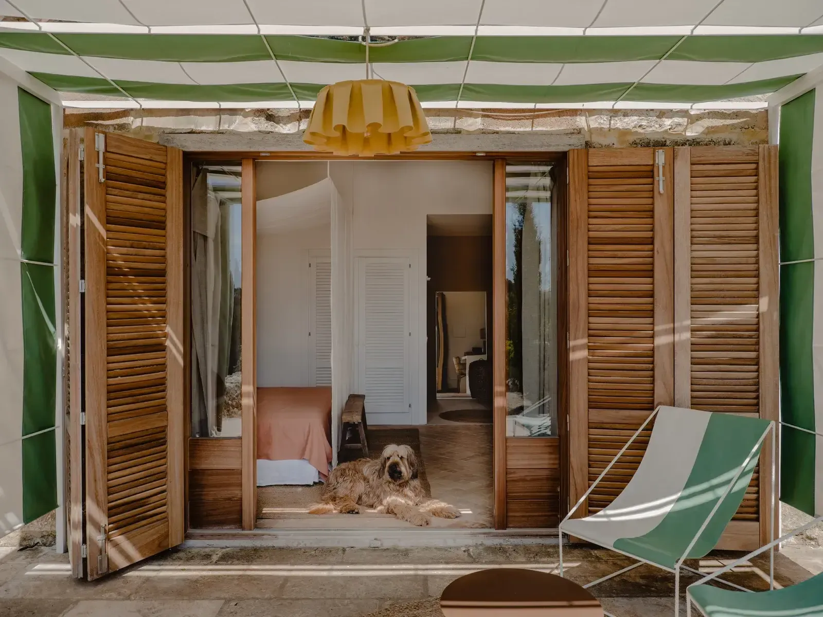 Amagatay, Balearic Islands — Open wooden louvered doors frame sunlit bedroom with terracotta floors, green striped canvas chair, and golden pendant light.
