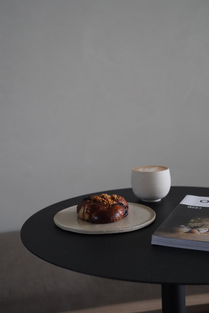 Hotel âme, Europe — Minimalist round black table with pastry on ceramic plate, cappuccino in white cup, and magazine, clean aesthetic lighting.