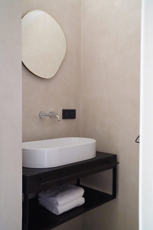Hotel âme, Europe — Modern minimalist bathroom with organic curved mirror, white vessel sink on black floating vanity, warm beige walls, and sculptural design details.