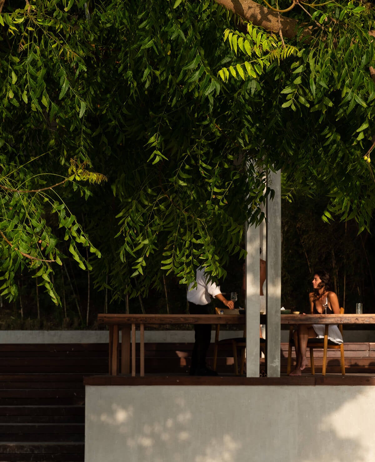 Anakampung, Asia — Tropical pavilion with suspended greenery, concrete and timber furnishings, dappled sunlight, contemporary hospitality design.