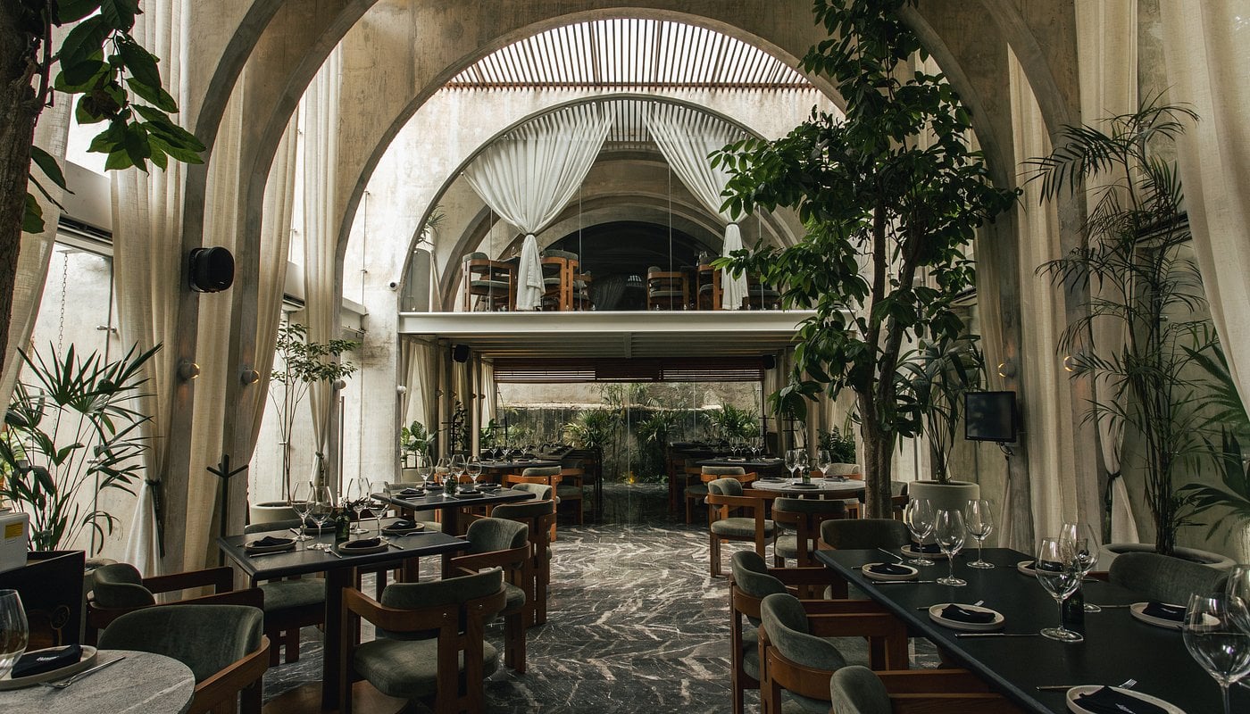 Ariadna, Mexico — Vaulted stone dining hall with soaring arches, natural light, potted plants, wood furniture, and garden views through floor-to-ceiling windows.