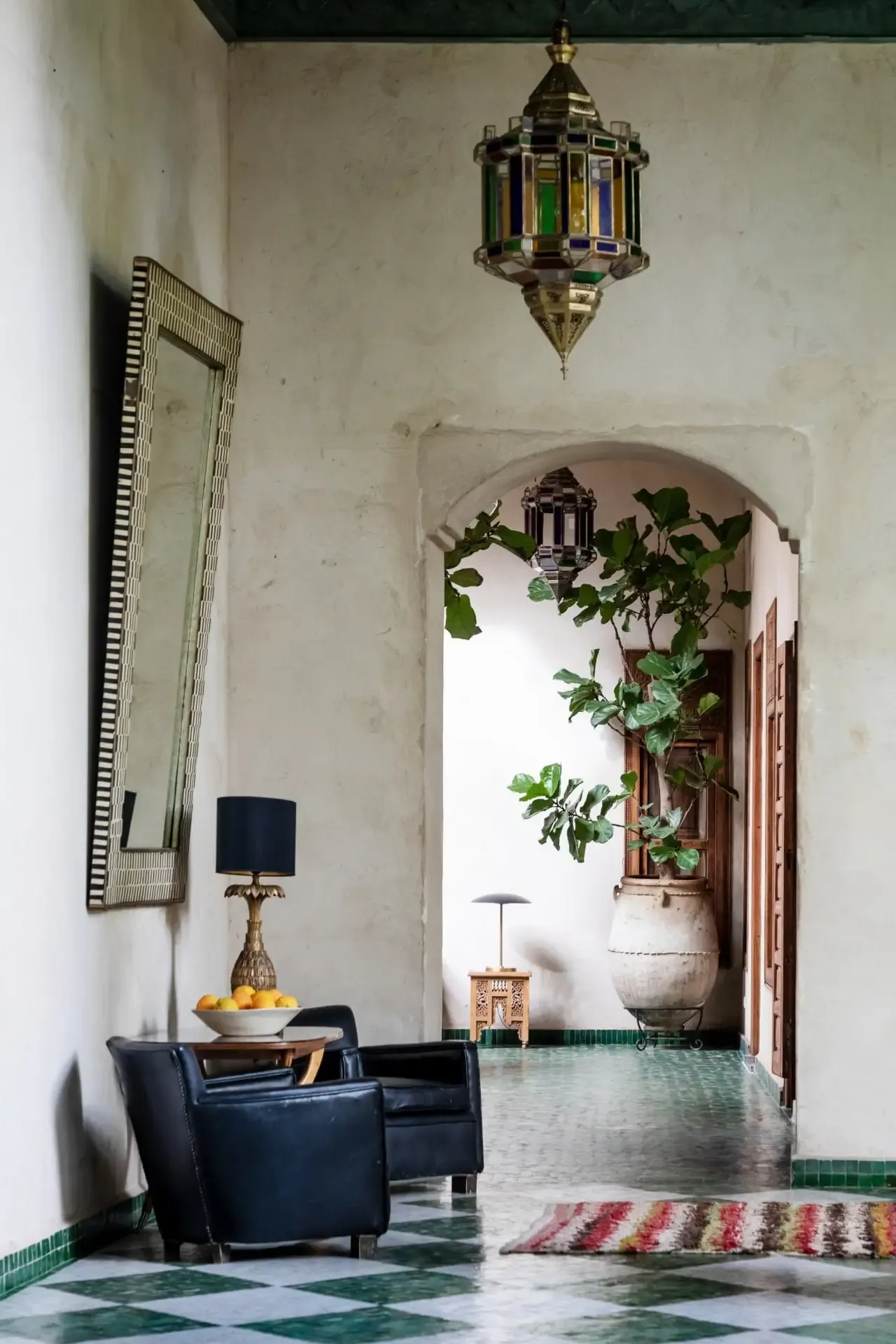 El Fenn, Marrakech, Morocco — Moroccan riad entry hall with zellige tile floor, brass lantern, arched doorway, potted climbing ivy, navy leather chair, and brass table lamp.