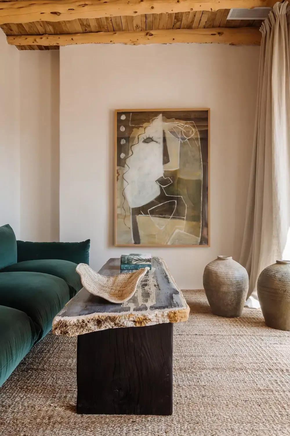 Finca La Inika, Balearic Islands, Spain — Minimalist living room with exposed wooden beams, emerald curved sofa, earthenware vessels, and abstract portrait above stone coffee table.