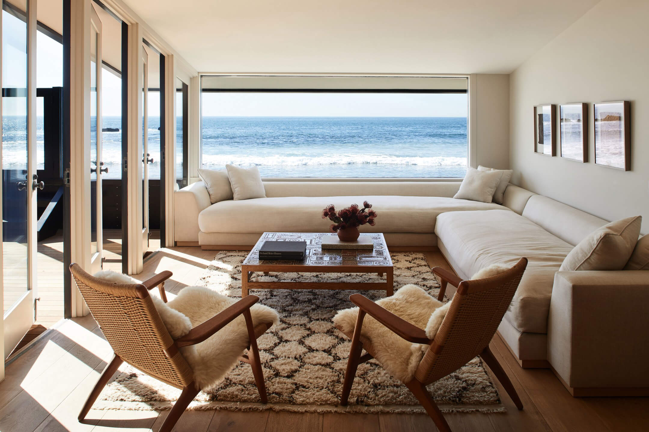 Jason Statham’s Malibu Beach House | sold — Oceanfront living room with curved cream sofa, mid-century wooden chairs, and expansive beach views through floor-to-ceiling windows.