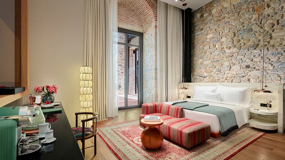 Aliée Istanbul, Istanbul, Turkey — Luxury hotel bedroom with exposed brick wall, striped red ottomans, brass pendant lighting, and floor-to-ceiling curtains opening to courtyard.
