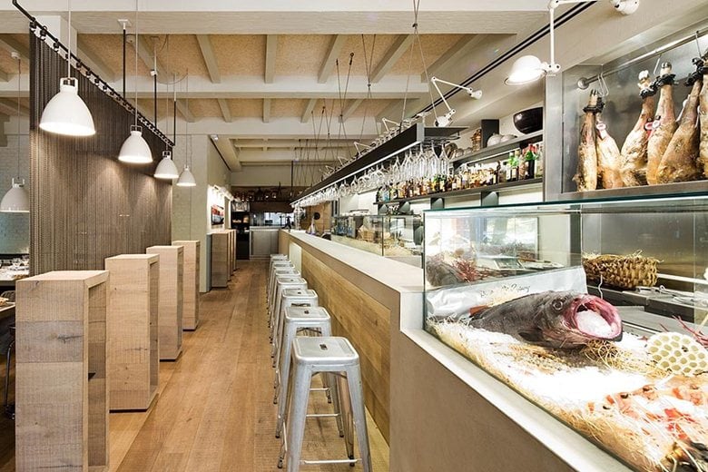 El Pescador, Europe — Modern seafood market with open kitchen, wooden display cases, hanging cured meats, marble counters, pendant lighting, and industrial-minimalist design.