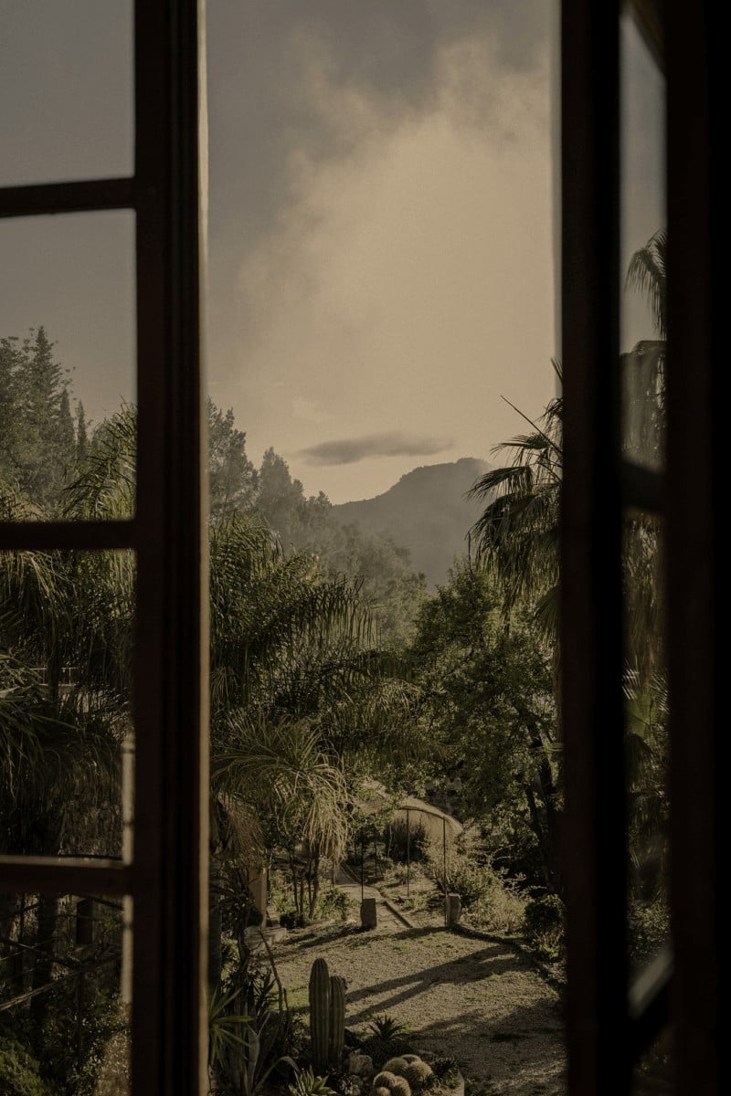 Hotel Corazón, Balearic Islands — View through dark wooden window frame to palm-lined courtyard, mountains, and clouded sky at dusk.