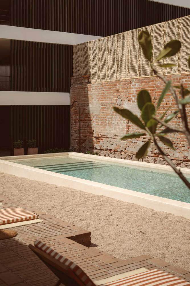 Baja Club, Mexico — Minimalist poolside courtyard with exposed brick wall, cactus, striped loungers, and modernist architecture in warm earth tones.