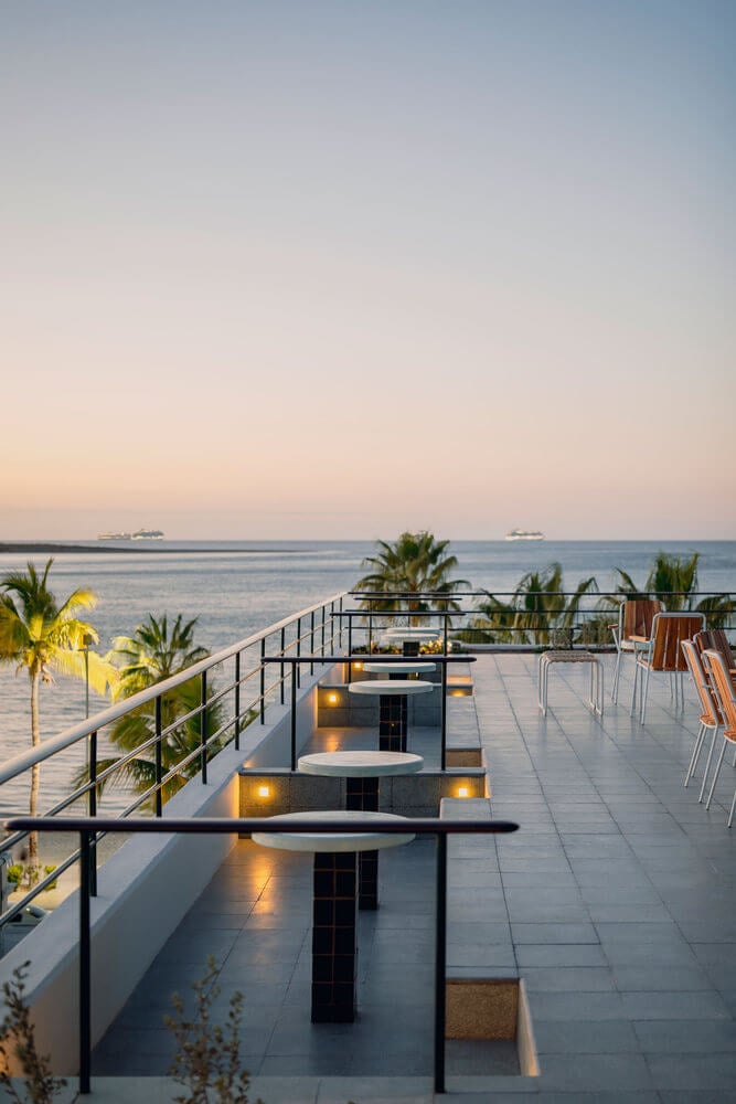 Rooftop Bar, Mexico — Modern terrace at dusk with illuminated tables, palm trees, Mediterranean Sea views, and anchored ships at horizon.