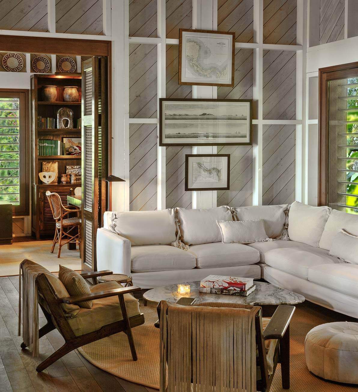 Barboa Bar, All Destinations — Diagonal wood-paneled living room with cream sectional, mid-century lounge chairs, marble coffee table, and framed coastal artwork beneath gridded white beams.