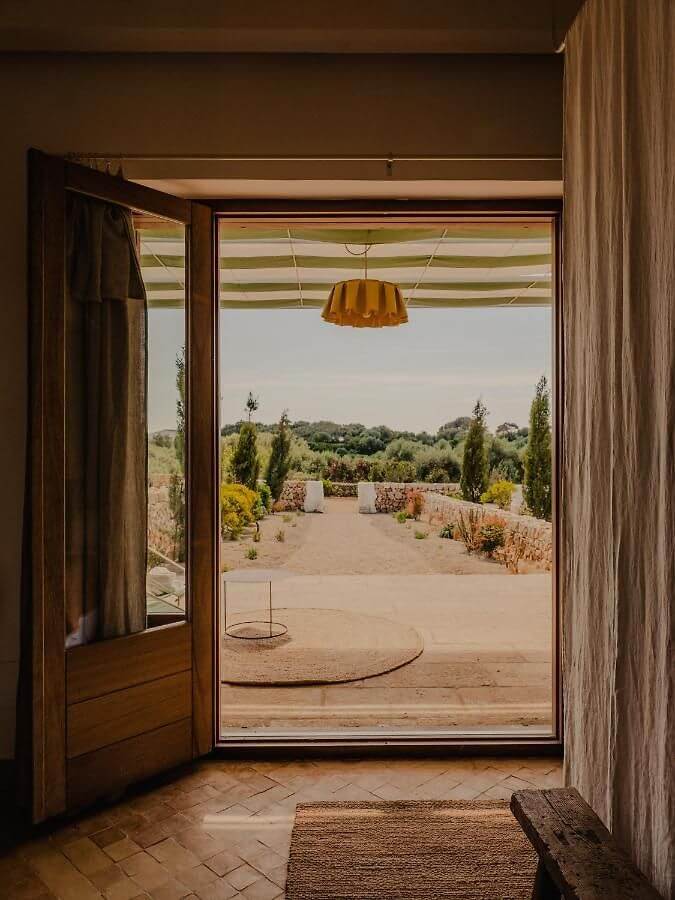 Amagatay, Balearic Islands — Open wooden door framing terracotta courtyard with striped awning, cypress trees, and Mediterranean landscape beyond.