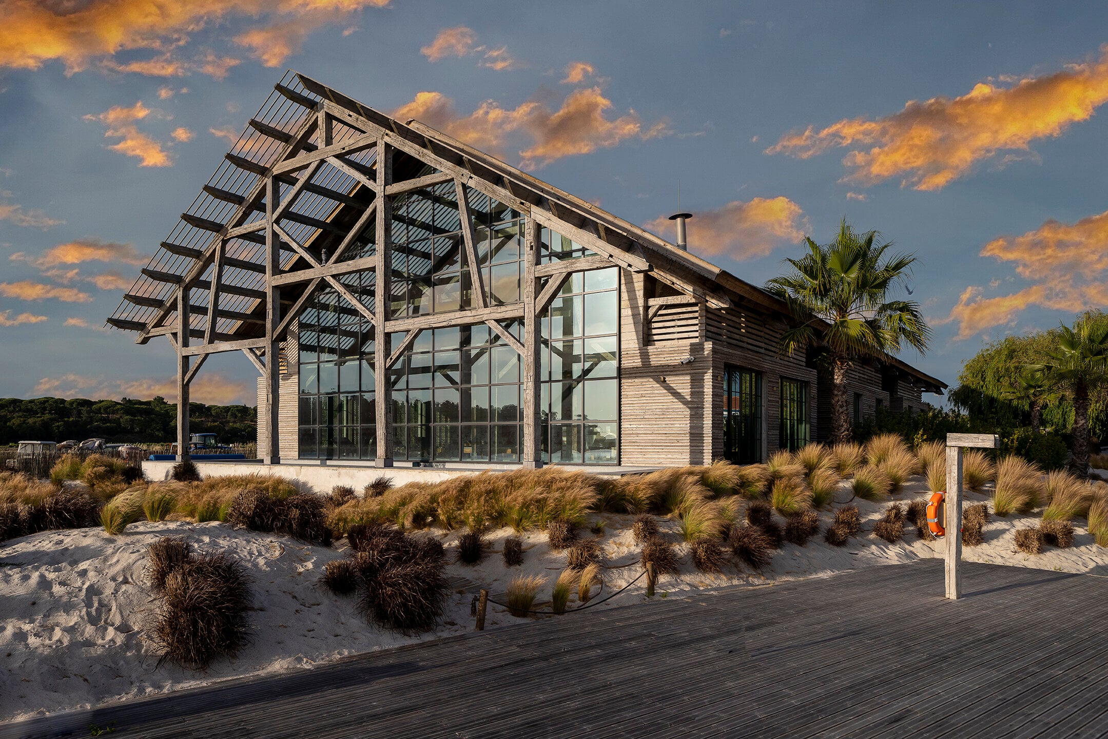 Quinta da Comporta, Portugal — Contemporary timber-frame beach pavilion with expansive glazing, weathered wood siding, and solar panels overlooking dunes and native coastal grasses at golden hour.