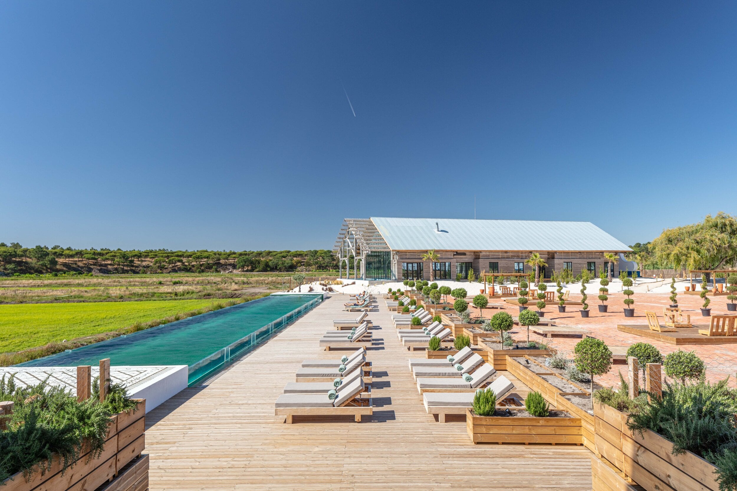 Quinta da Comporta, Portugal — Wooden deck with loungers overlooking turquoise infinity pool, modernist barn architecture, and marshland landscape under clear sky.