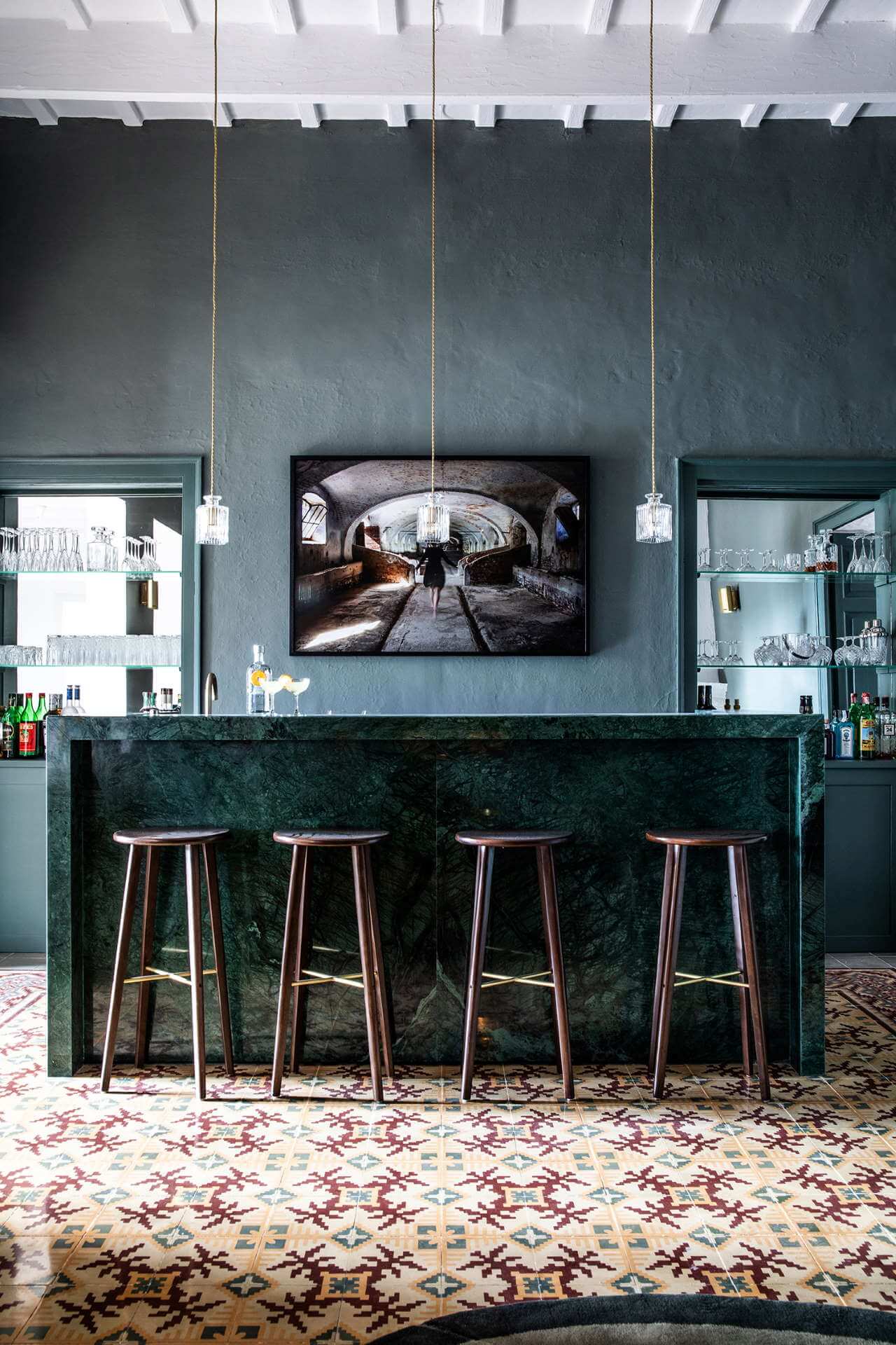 Es Bec d’Aguila — Es Bec d'Aguila, Balearic Islands, Spain — Dark green marble bar with wooden stools beneath concrete walls, pendant lights, and framed tunnel photograph, geometric tile flooring.