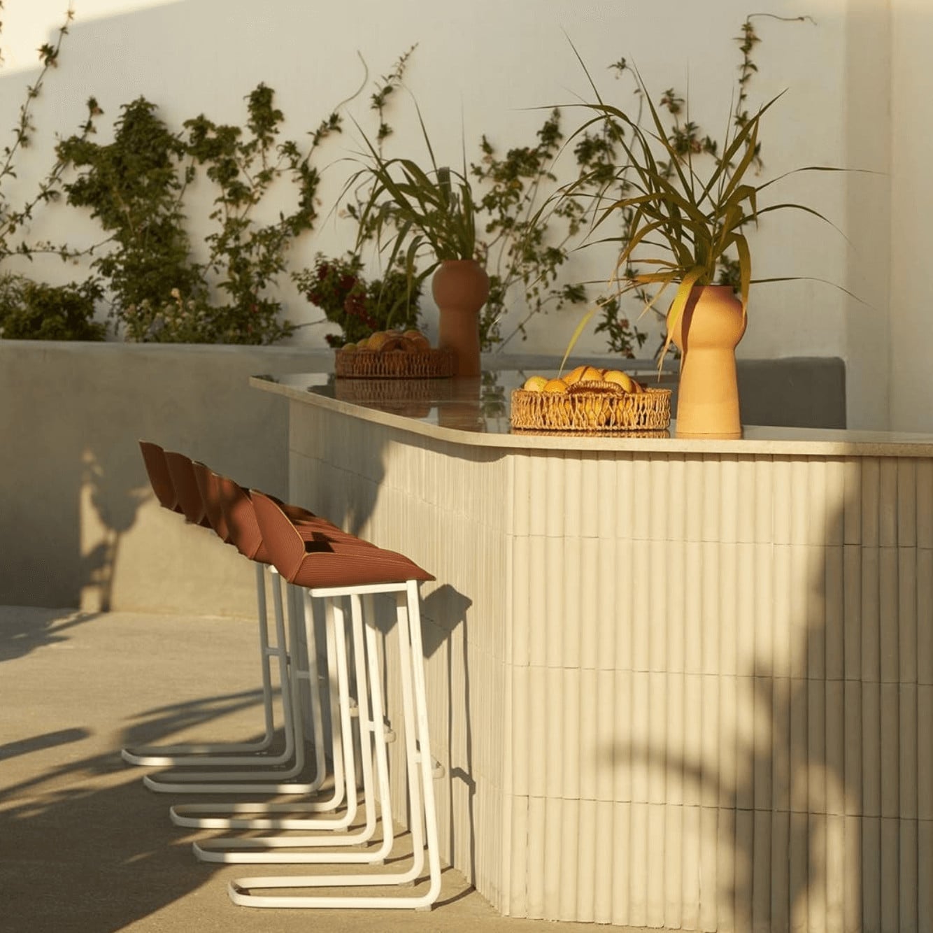 Pārocks, Europe — Minimalist outdoor bar with ribbed white cabinetry, terracotta stools, dried botanicals, warm afternoon light, and geometric shadows.