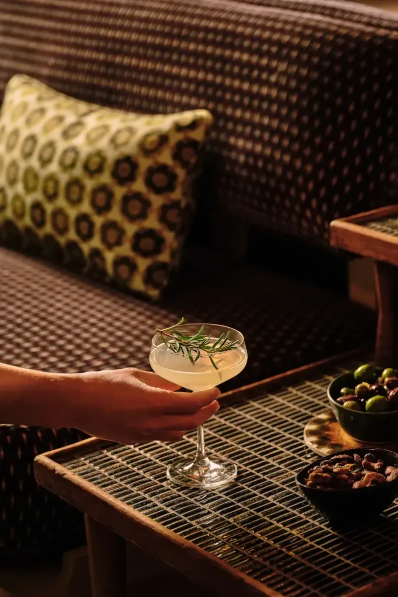 Dew Bar, North America — Hand holding rosemary-garnished cocktail on woven table with olives and nuts, rattan pillow and textured wall backdrop.