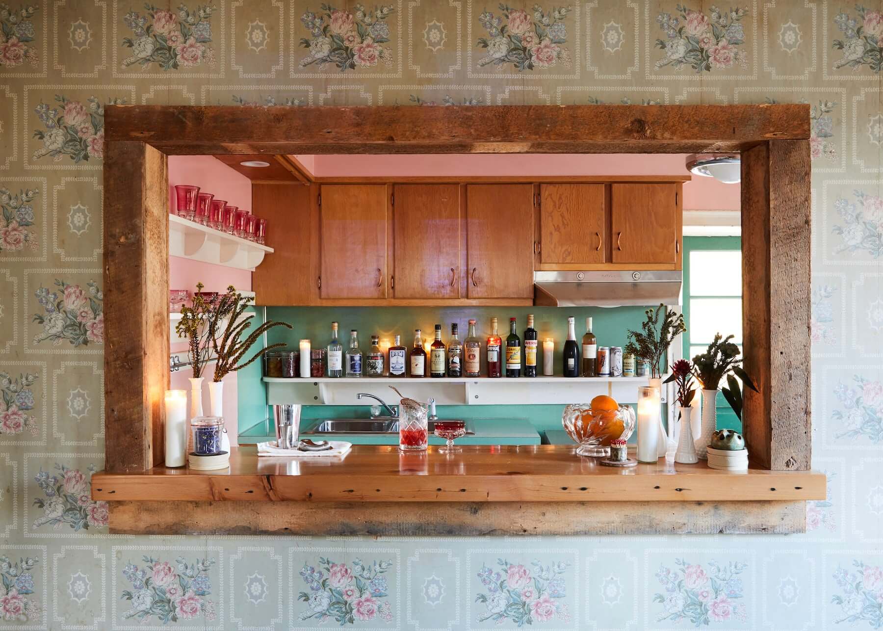 The Airport Rooms, North America — Recessed wooden bar with turquoise backsplash, vintage floral wallpaper, spirits collection, candlelit shelving, botanical arrangements, mid-century hospitality design.