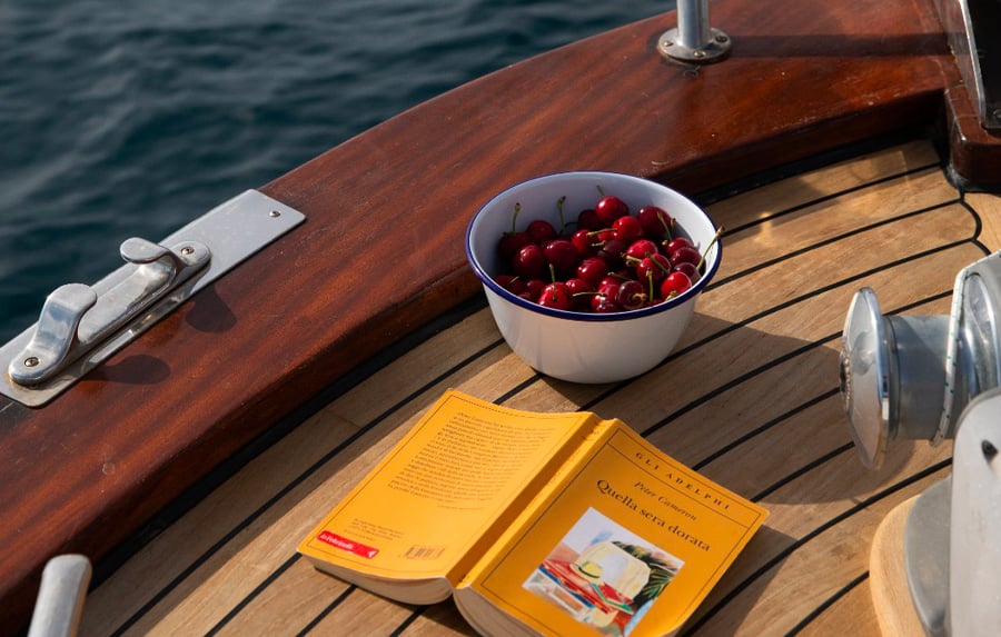 Boat Margherita Moroseta, Europe — Open wooden deck with fresh cherries in enamel bowl, yellow book, and maritime hardware in sunlight.
