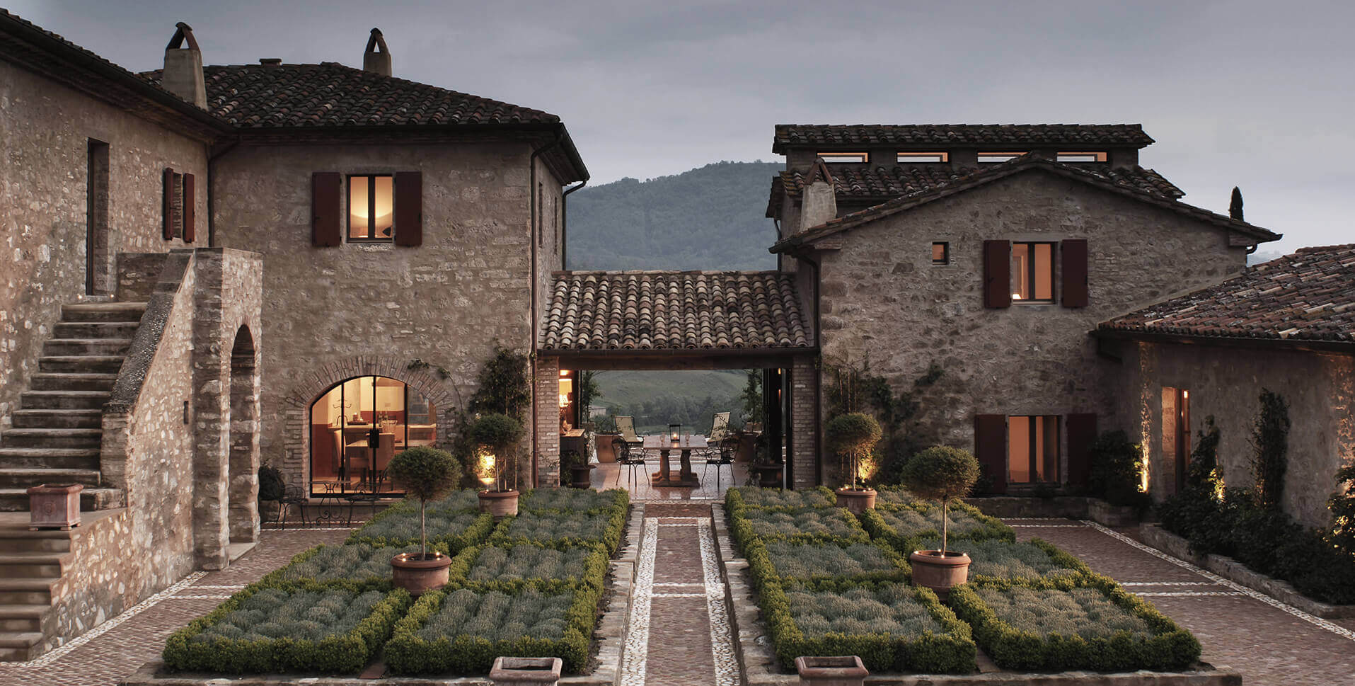 Castello di Reschio, Italy — Tuscan stone courtyard with arched loggia, lavender gardens, terracotta planters, and illuminated interiors at dusk.