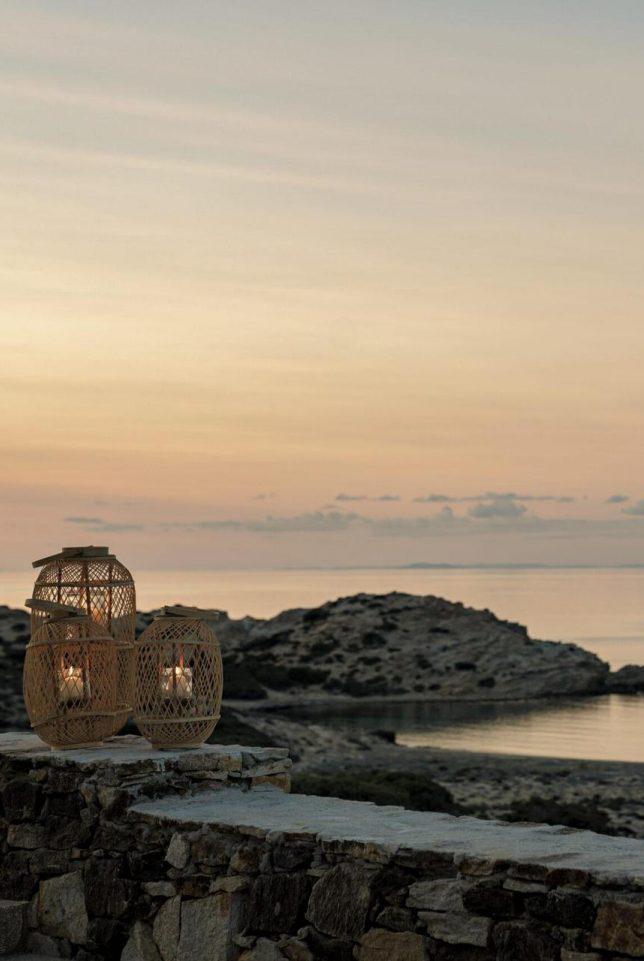 The Rooster, Europe — Woven rattan lanterns with candlelight on rocky coastal ledge at dusk, calm sea and distant islands under soft peachy sky.