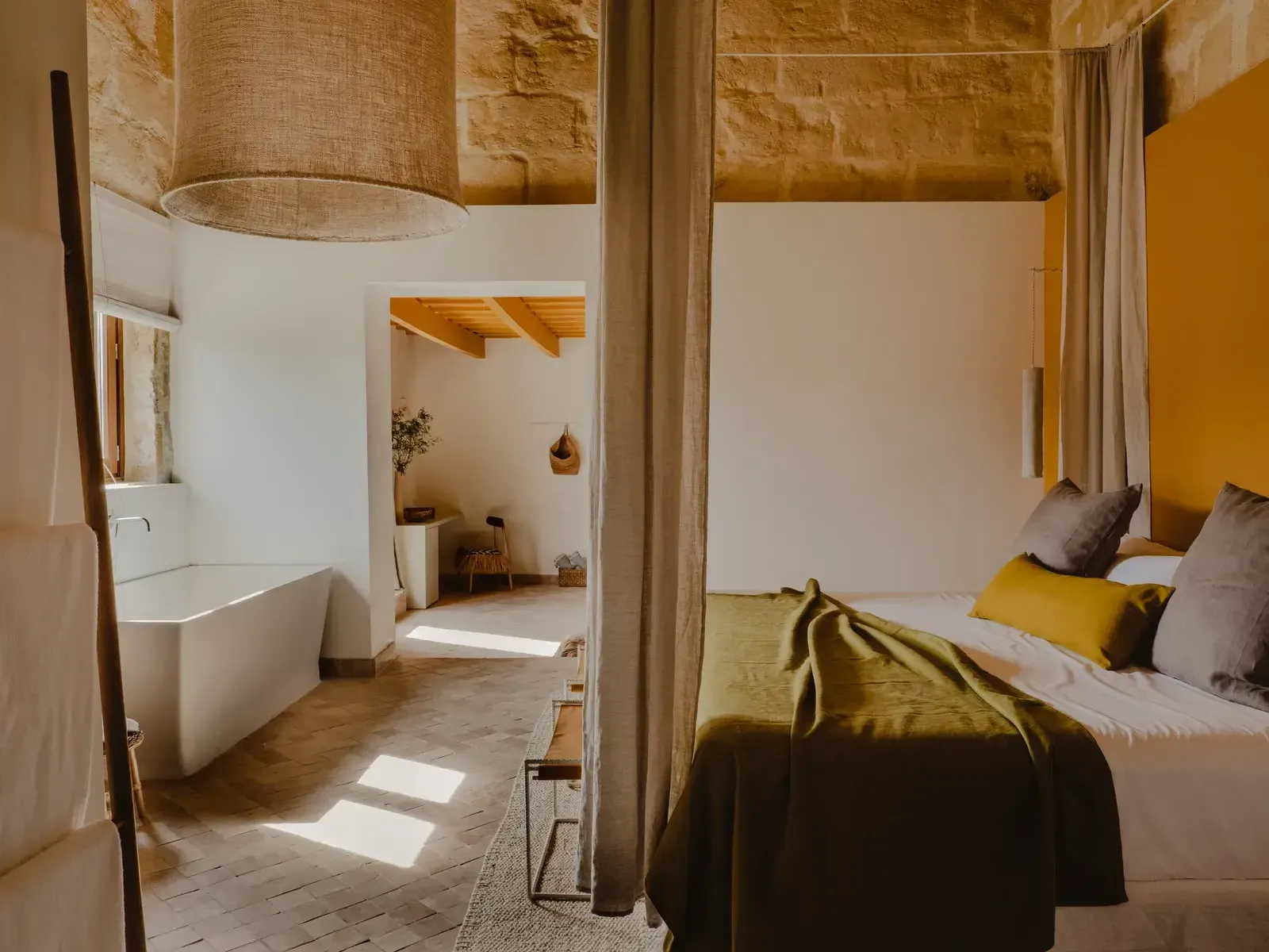 Amagatay, Balearic Islands — Minimalist bedroom-bathroom suite with ochre accents, natural light, stone walls, linen furnishings, and open-plan luxury design.
