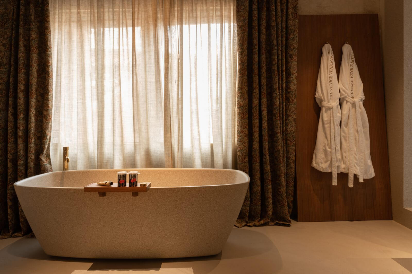 The Venice Venice Hotel, Europe — Freestanding oval stone bathtub with brass tray beside sheer and heavy bronze curtains filtering natural light, two white robes hanging on warm brown wall.