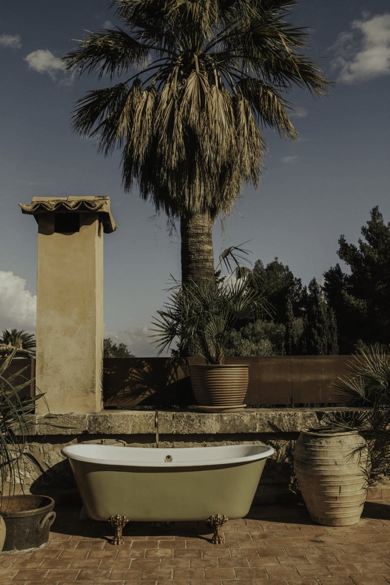 Hotel Corazón, Balearic Islands — Freestanding bathtub on terrace with palm tree, stone chimney, and Mediterranean landscape under clear sky.