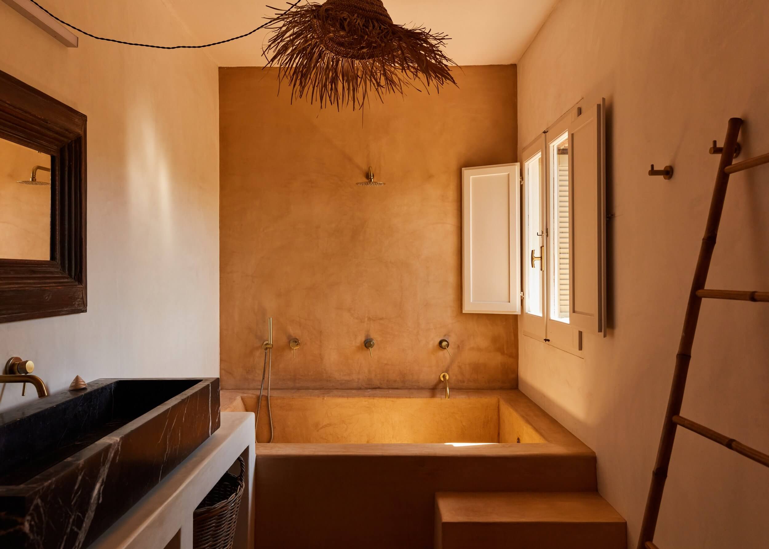 Parco Dei Sesi, Europe — Ochre plaster bathroom with sunken soaking tub, woven pendant light, wooden ladder towel rack, and minimalist bronze fixtures.