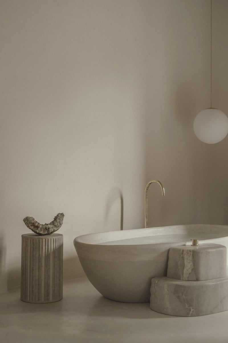 Hotel Corazón, Balearic Islands, Spain — Minimalist bathroom with sculptural stone bathtub, travertine fixtures, and curated wellness accessories against neutral walls.