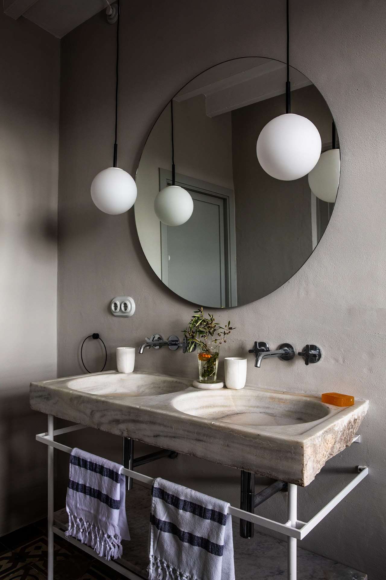 Es Bec d’Aguila — Es Bec d'Aguila, Balearic Islands, Spain — Minimalist bathroom with dual marble basins, round mirror, and pendant globe lights above stone countertop.