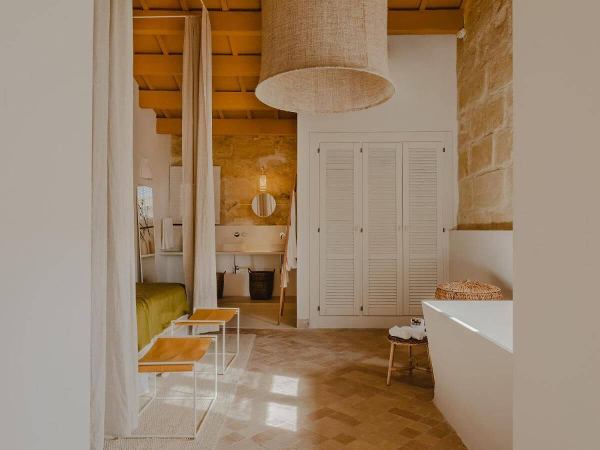 Amagatay, Balearic Islands — Minimalist hotel corridor with ochre stone walls, linen pendant light, white louvered wardrobes, and warm terracotta flooring.