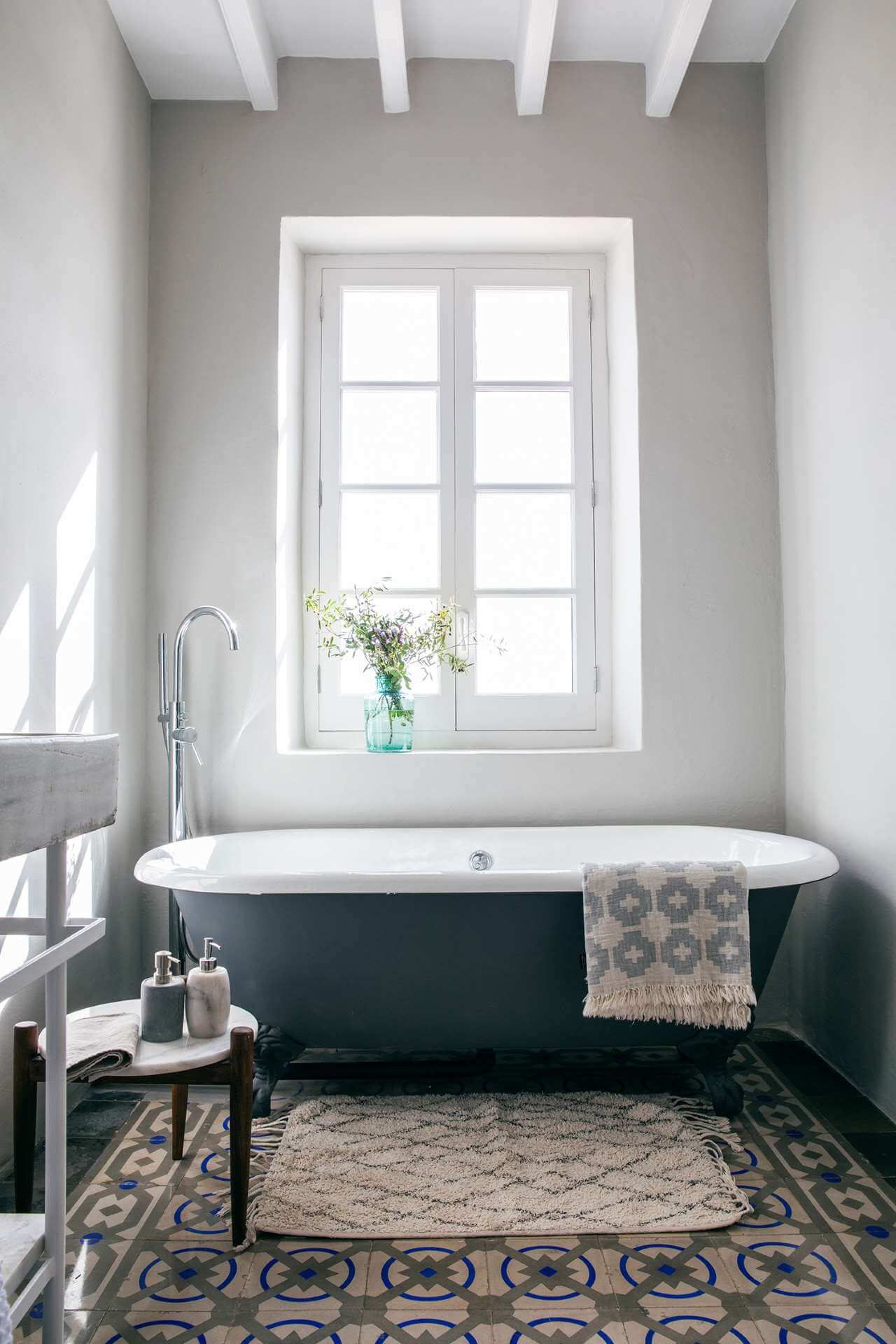 Es Bec d’Aguila — Es Bec d'Aguila, Balearic Islands — Black freestanding bathtub with white interior beneath tall casement window, geometric patterned tile floor, natural light, minimalist bathroom interior.
