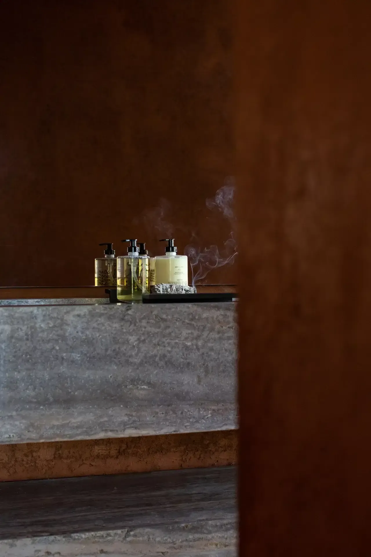 Further Hotel, Asia — Concrete counter with amber glass pump bottles and lit candle against warm terracotta wall, minimalist luxury spa aesthetic.