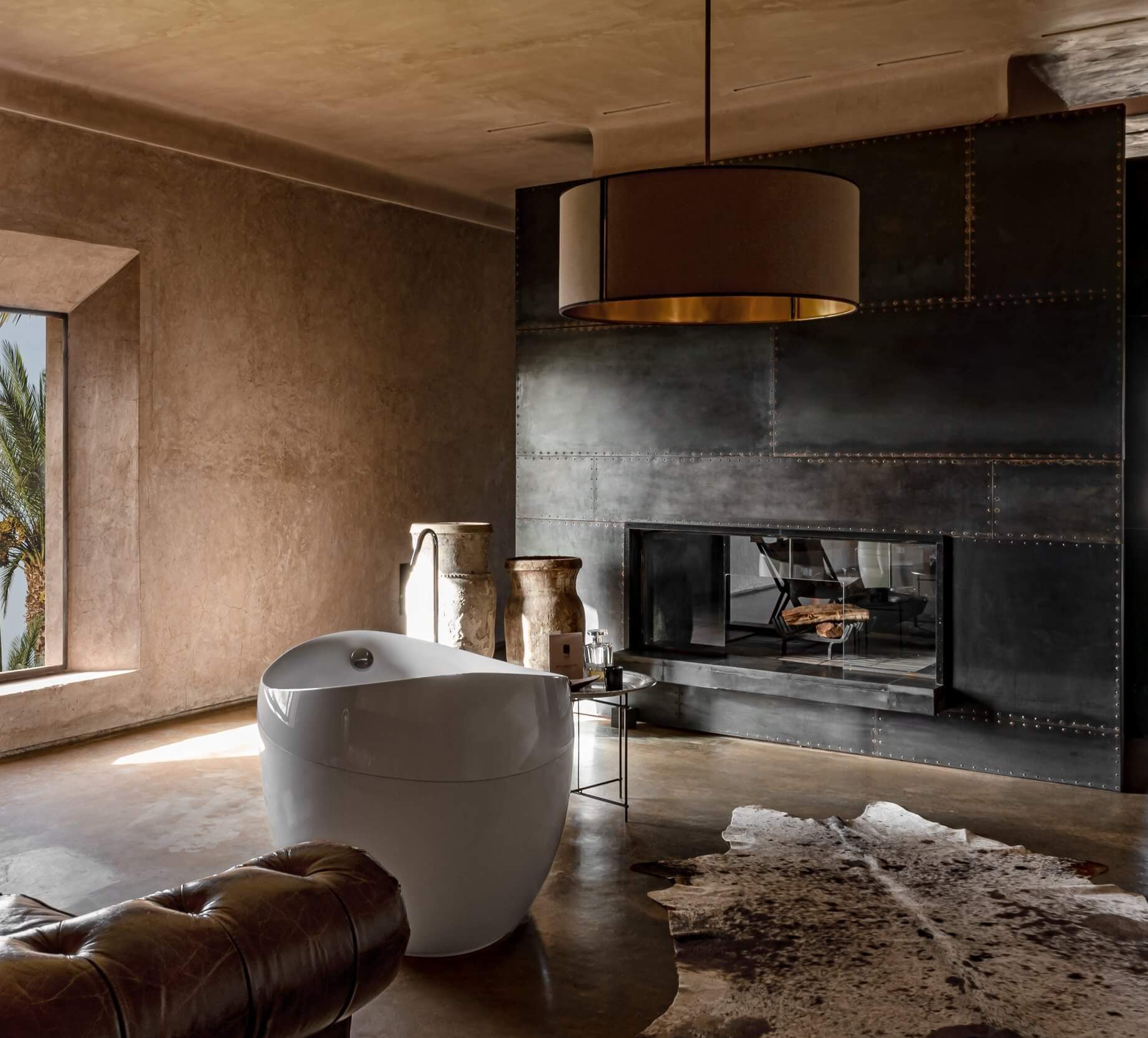 Villa D, Africa — Minimalist bathroom with sculptural white soaking tub, industrial metal fireplace, warm plaster walls, brass pendant light, natural stone flooring, and curated décor.
