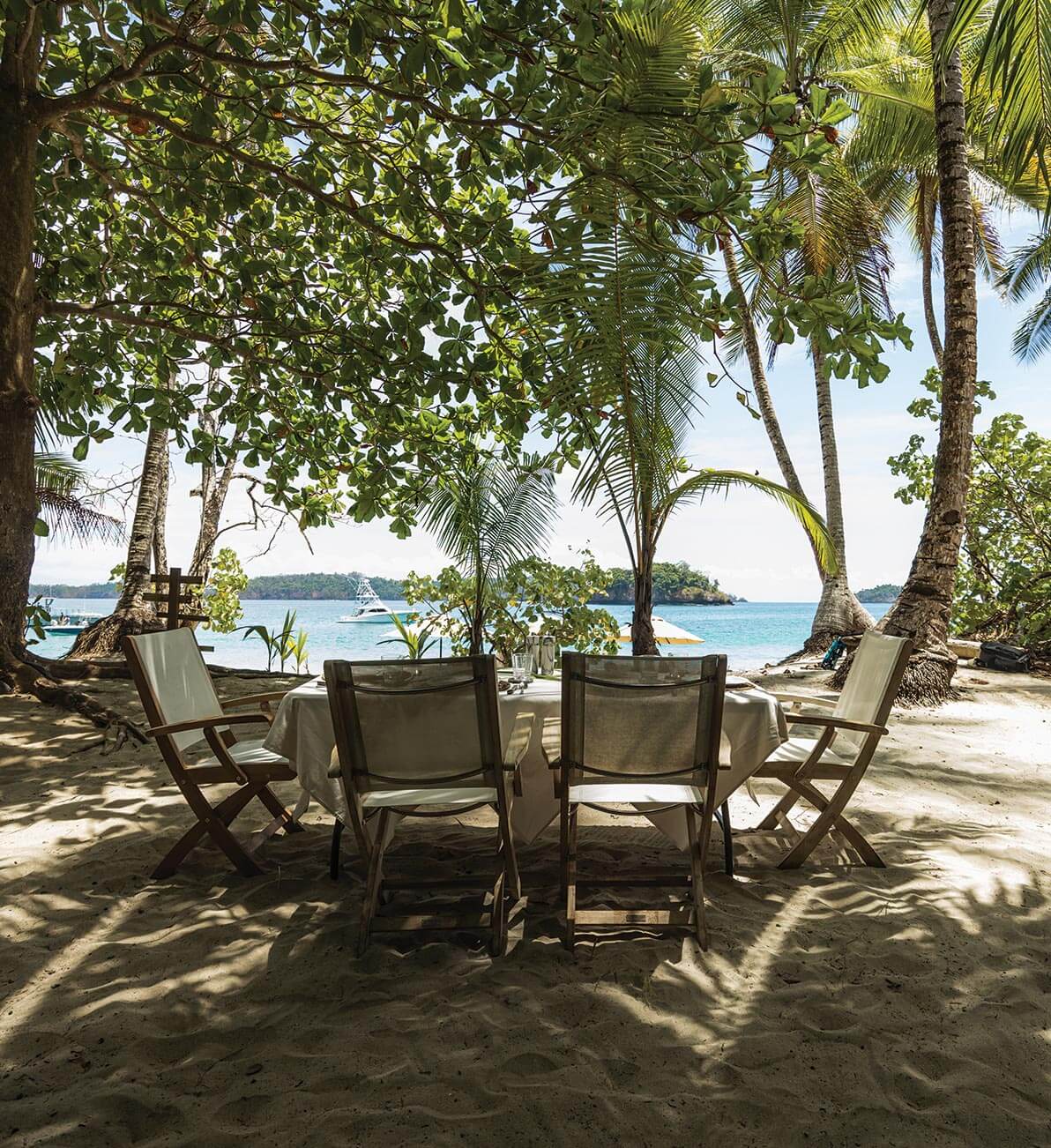 Beach BBQ, All Destinations — Teak lounge chairs arranged on sand beneath palm trees and tropical foliage, turquoise water and sailboat visible through opening.