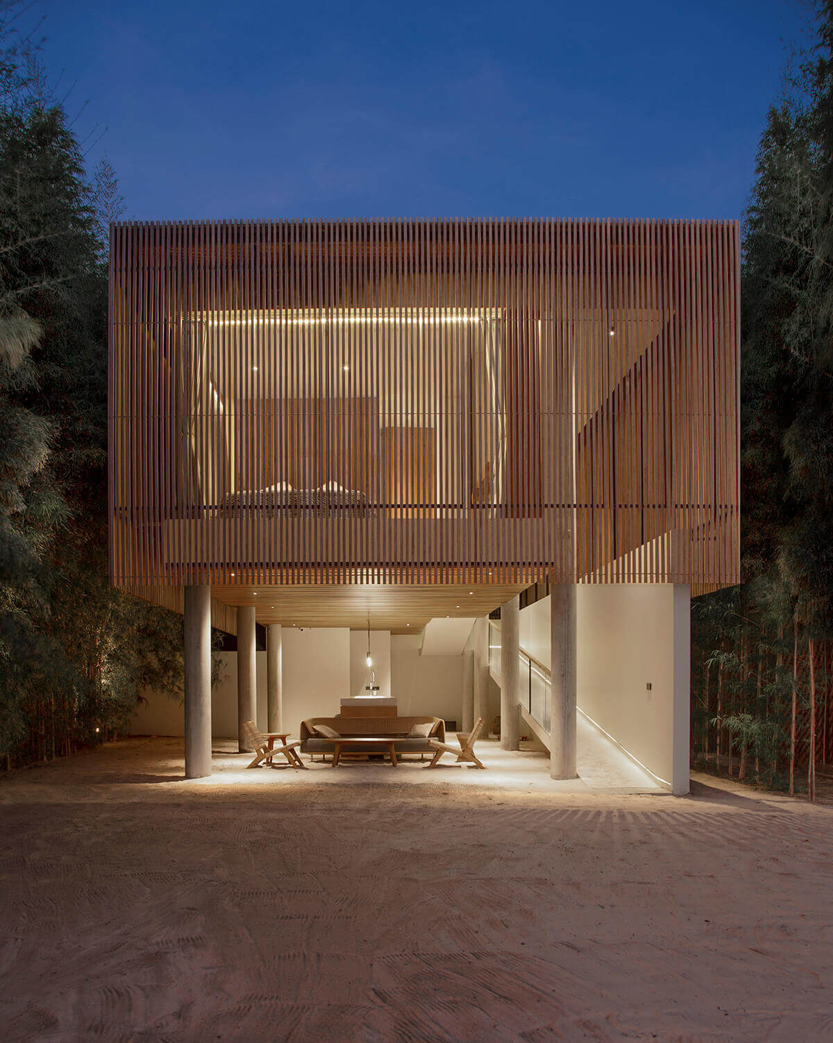 Innit Lombok, Asia — Modernist villa with oxidized copper louver facade, illuminated interior, open-air dining pavilion, and minimal concrete supports on sand at dusk.