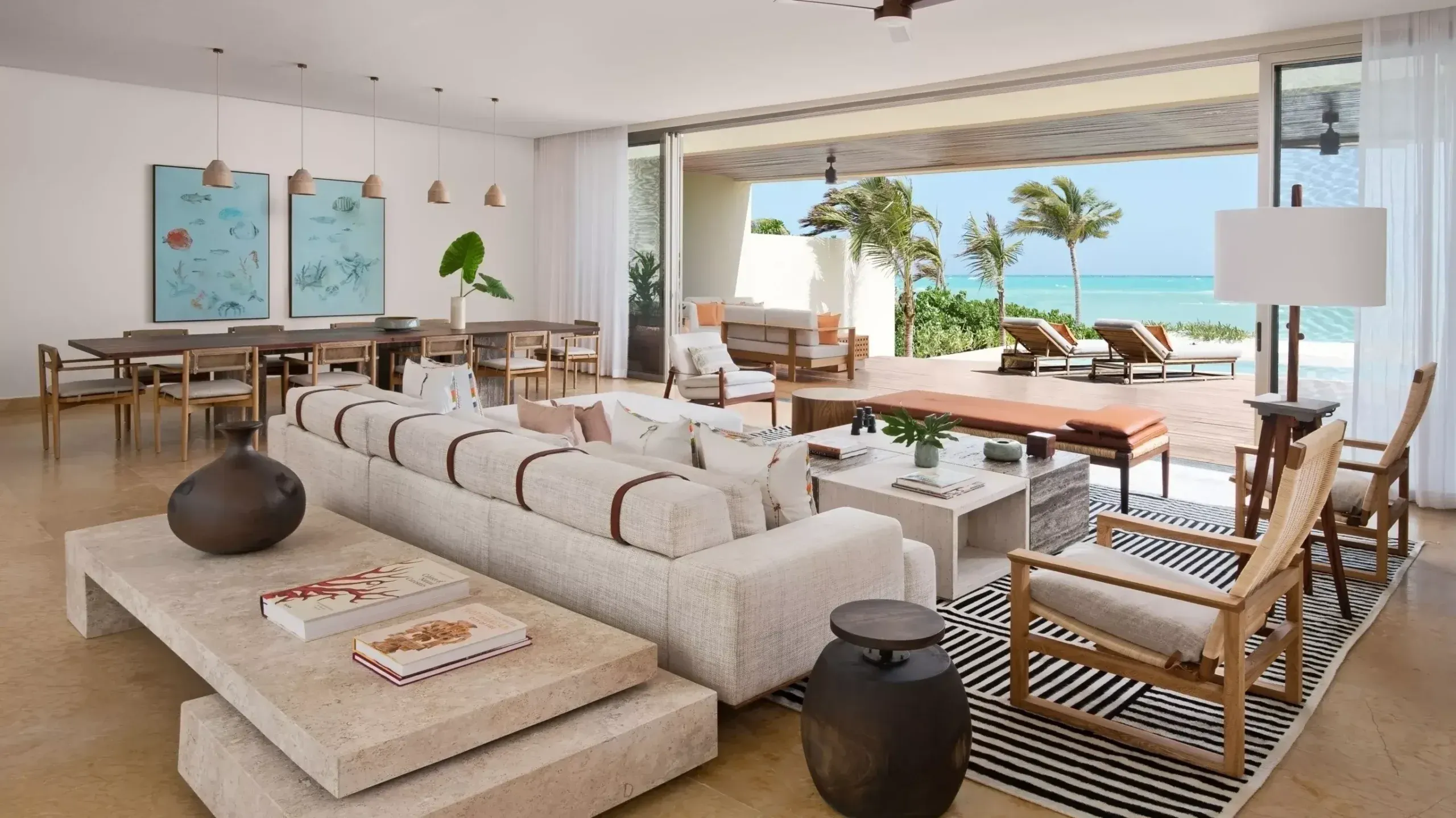 Rosewood Mayakoba, Mexico — Beachfront living room with curved cream sofa, wooden furniture, striped rug, ocean views, and palm trees at sunset.