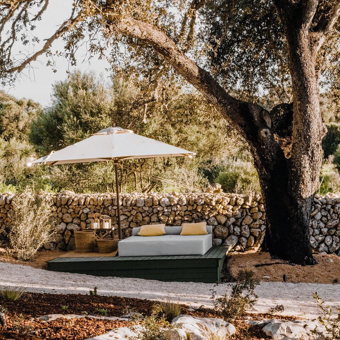 Amagatay, Balearic Islands — Outdoor daybed beneath ancient olive tree with cream umbrella, stone wall, and Mediterranean garden setting.