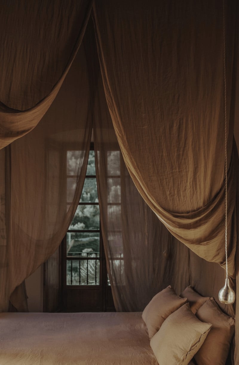 Hotel Corazón, Balearic Islands — Canopied bed with sheer brown linen curtains framing tall windows overlooking landscape and distant hills, soft natural light filtering through fabric.