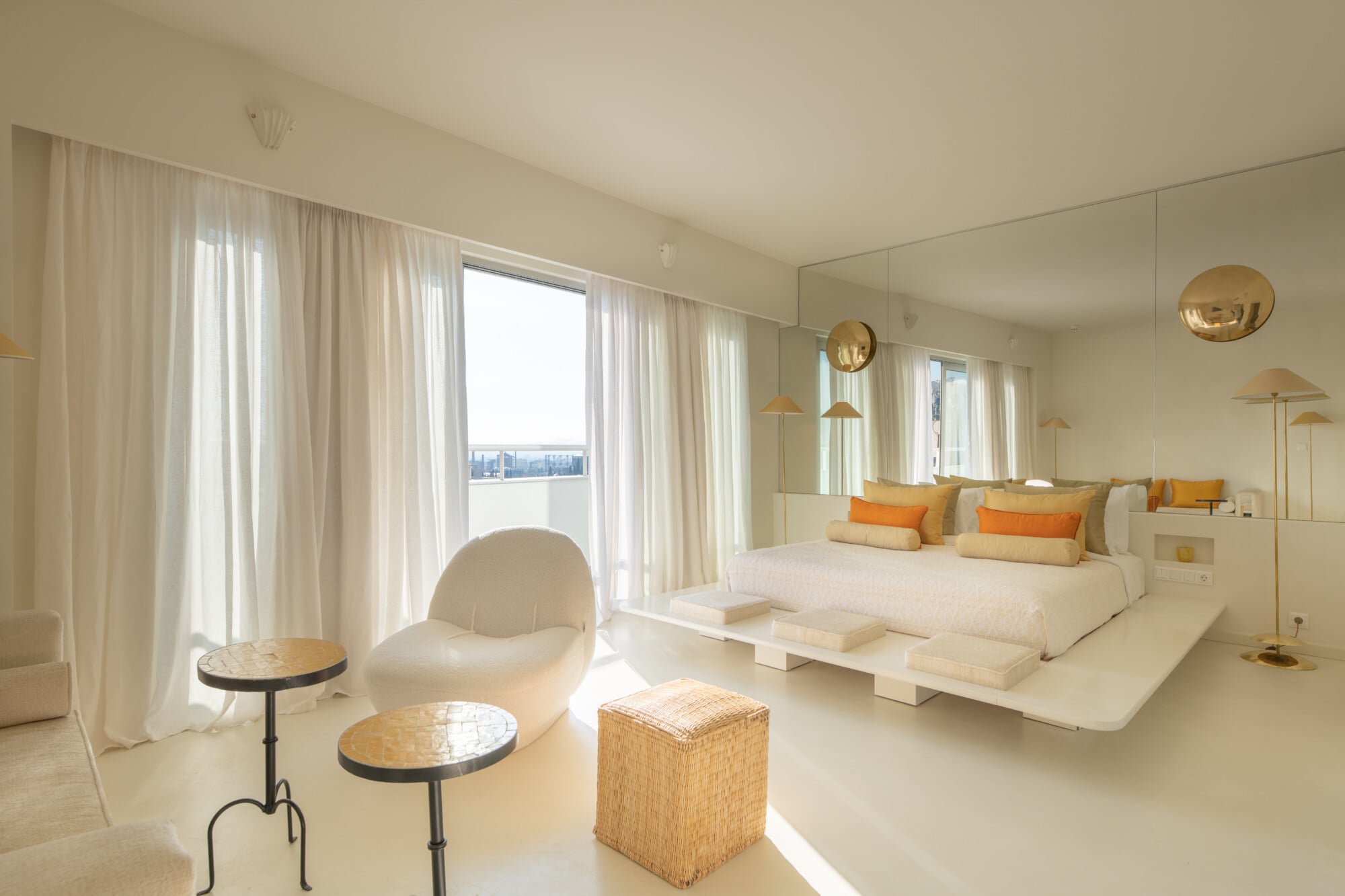 Apollo Palm, Europe — Minimalist bedroom suite with cream terrazzo flooring, floor-to-ceiling windows, brass pendant lights, and panoramic city views through sheer curtains.