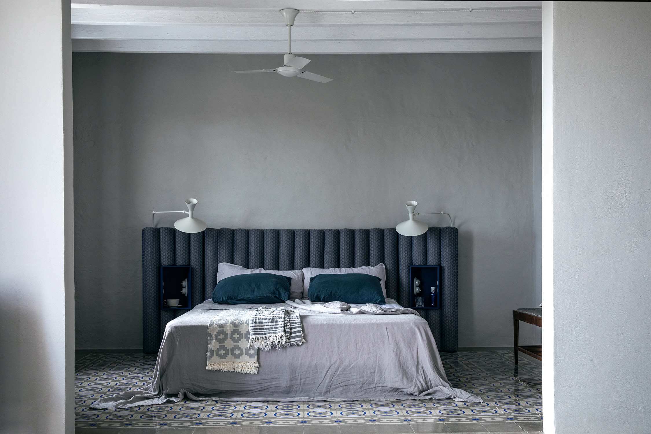 Es Bec d’Aguila — Es Bec d'Aguila, Balearic Islands, Spain — Minimalist bedroom with charcoal fluted headboard, white bedding, dual pendant lights, ceiling fan, geometric tile flooring, and neutral walls.
