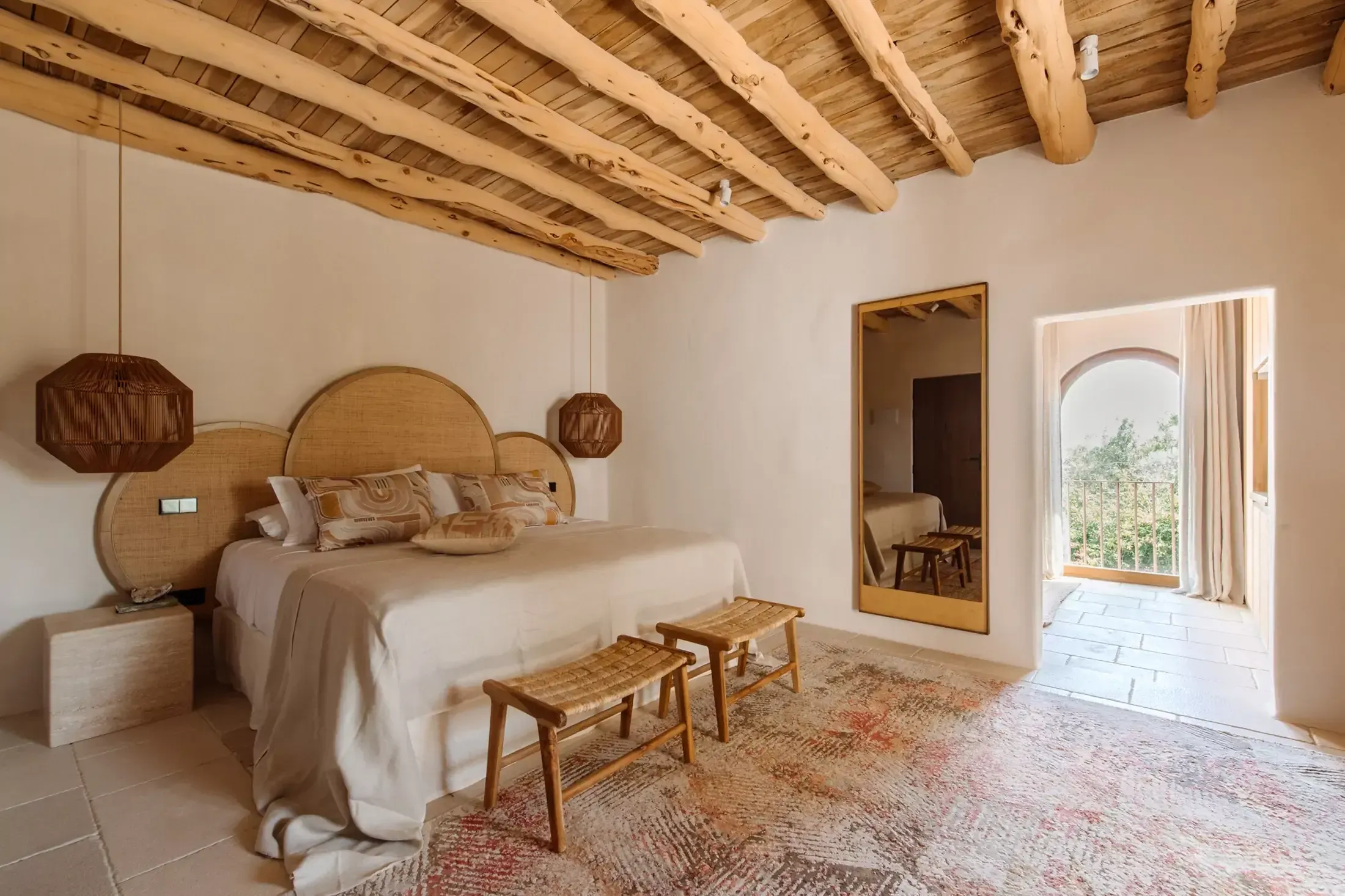 Finca La Inika, Balearic Islands — Minimalist bedroom with exposed wooden beams, woven rattan headboard, linen bedding, woven benches, and arched doorway opening to terrace.