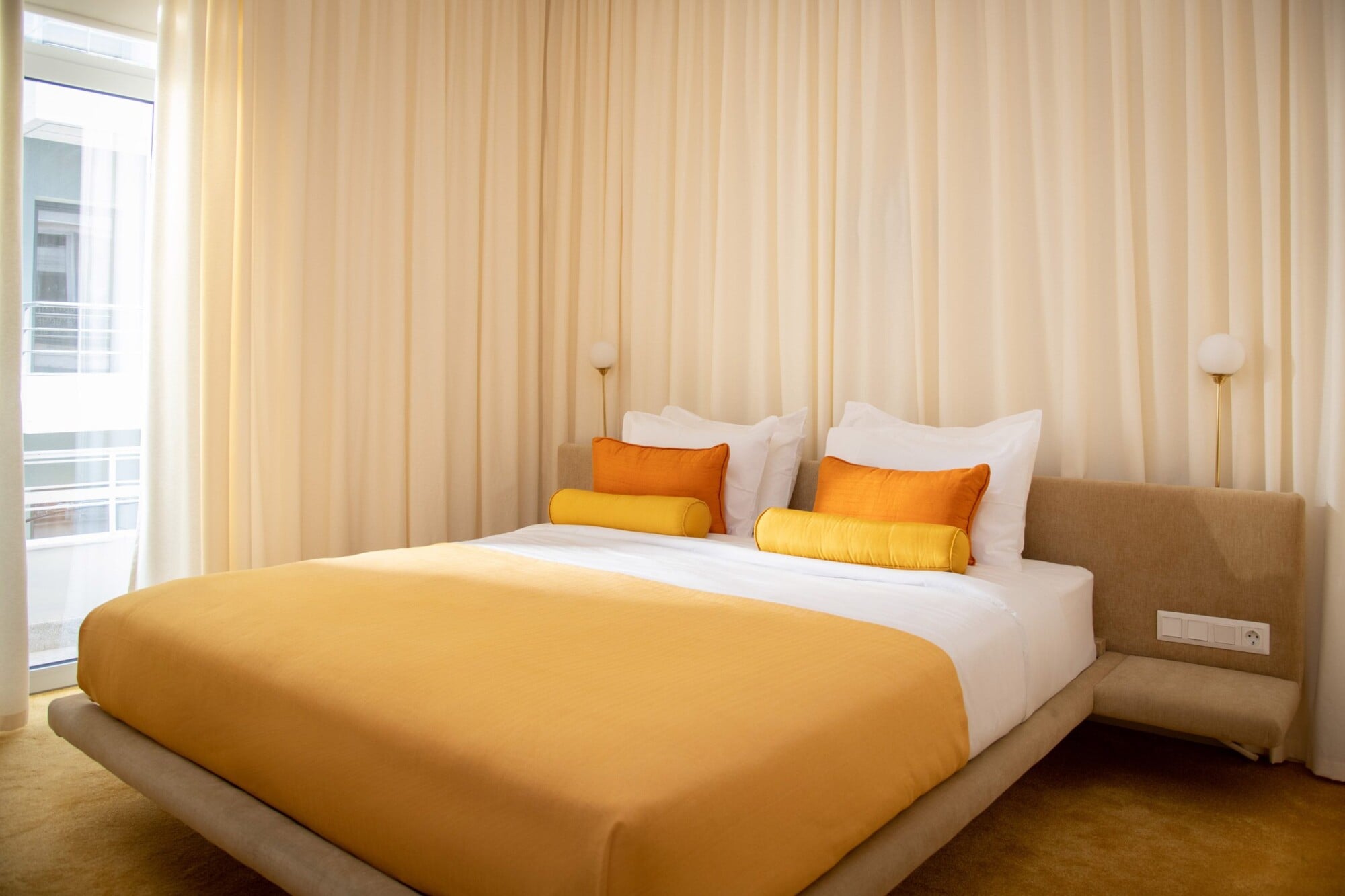 Apollo Palm, Europe — Modern hotel bedroom with golden yellow bedcover, cream upholstered headboard, orange accent pillows, and floor-to-ceiling sheer curtains creating warm natural light.