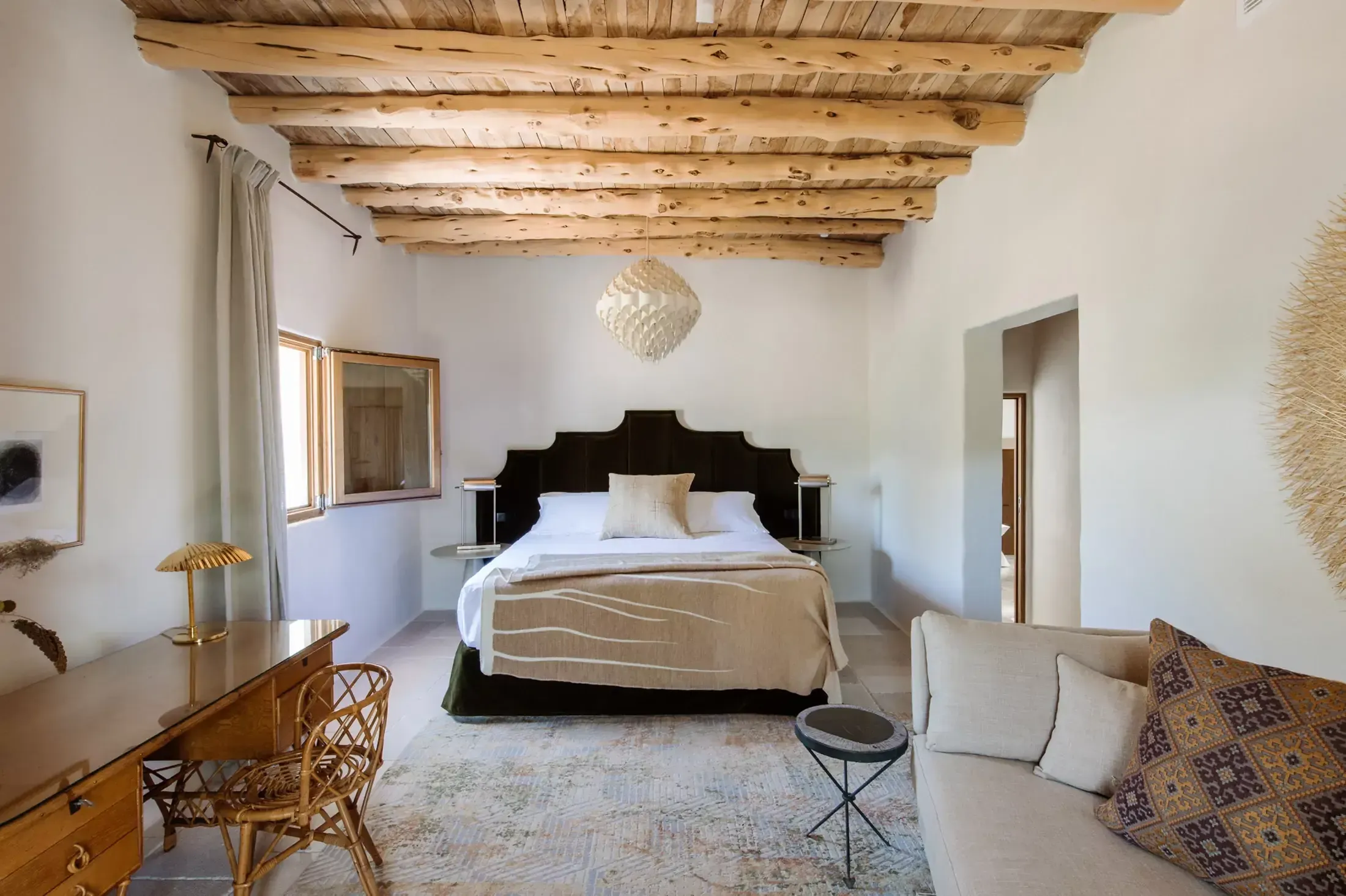Finca La Inika, Balearic Islands, Spain — Bedroom with black headboard, exposed wooden beams, white walls, pendant light, and minimalist furnishings in warm neutral tones.